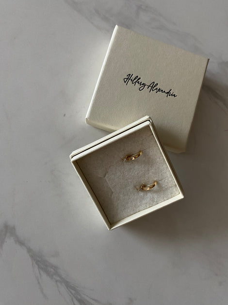 Two gold rings in a box with 'Hilary Alexander' branding on a marble surface.