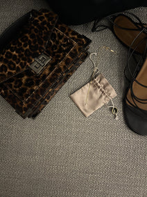 Leopard print clutch, Hillary Alexandria fine jewelry Vi necklace with lab diamond, and black sandals on a textured surface