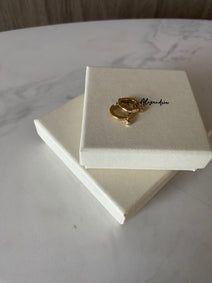 Gold earrings on a white jewelry box with 'Hillary Alexandria' branding on a marble surface.